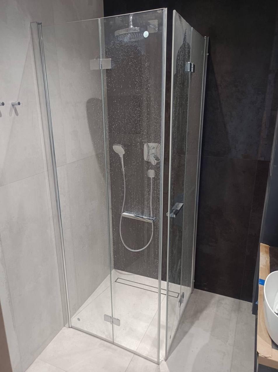 Szklana kabina prysznicowa z deszczownicą i odpływem liniowym, wykończona szarymi płytkami. Widoczne detale armatury i minimalistyczny design.