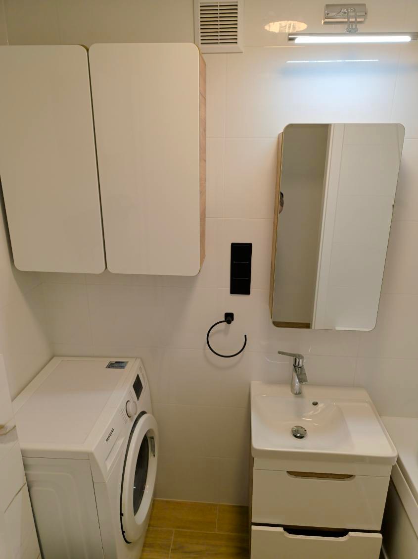 Jasna łazienka po remoncie: białe płytki, szafki z drewnianymi akcentami, pralka i umywalka z lustrem. Minimalistyczny design, czarne detale (wieszak, włącznik).