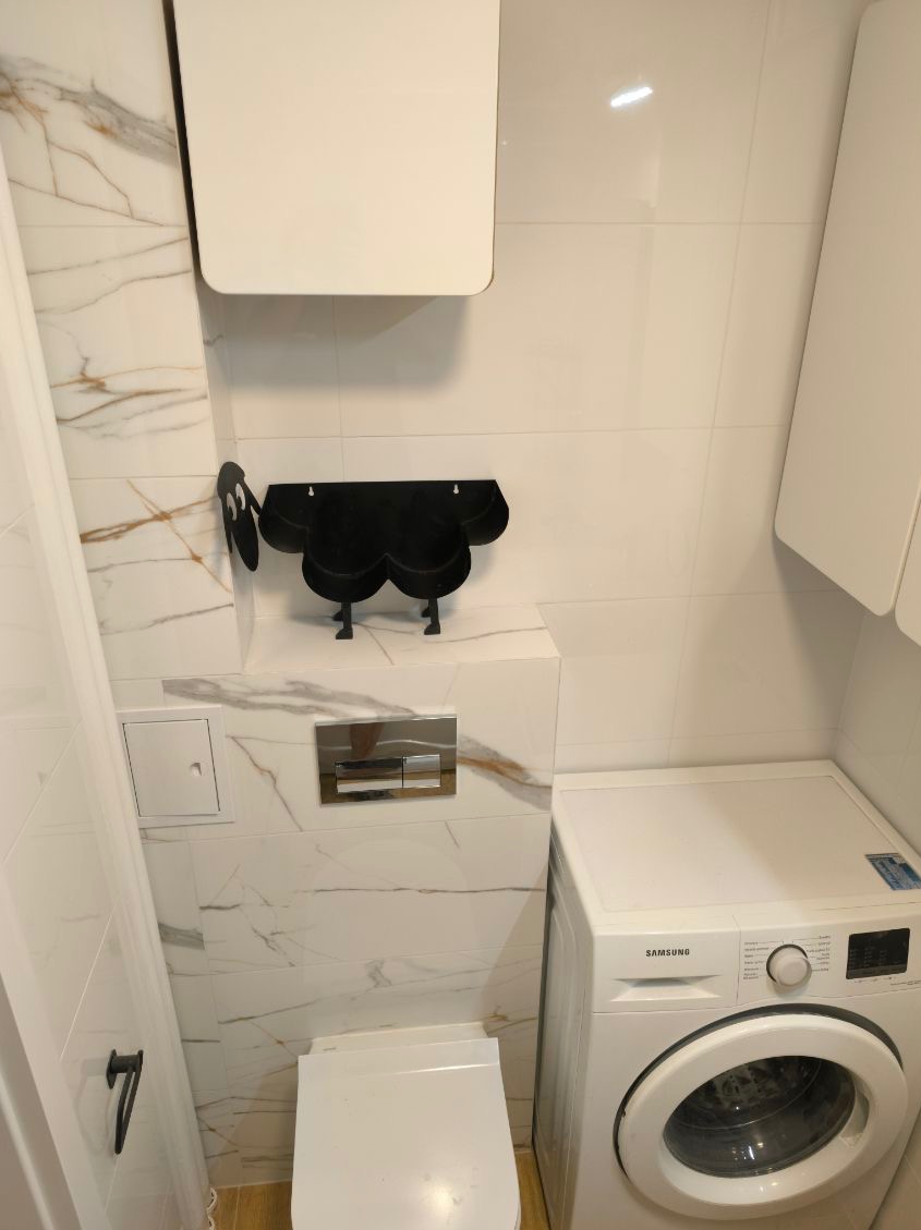 Biała łazienka z marmurowymi płytkami, wiszącą toaletą, pralką Samsung i czarnym, dekoracyjnym wieszakiem na ręczniki w kształcie chmurki.