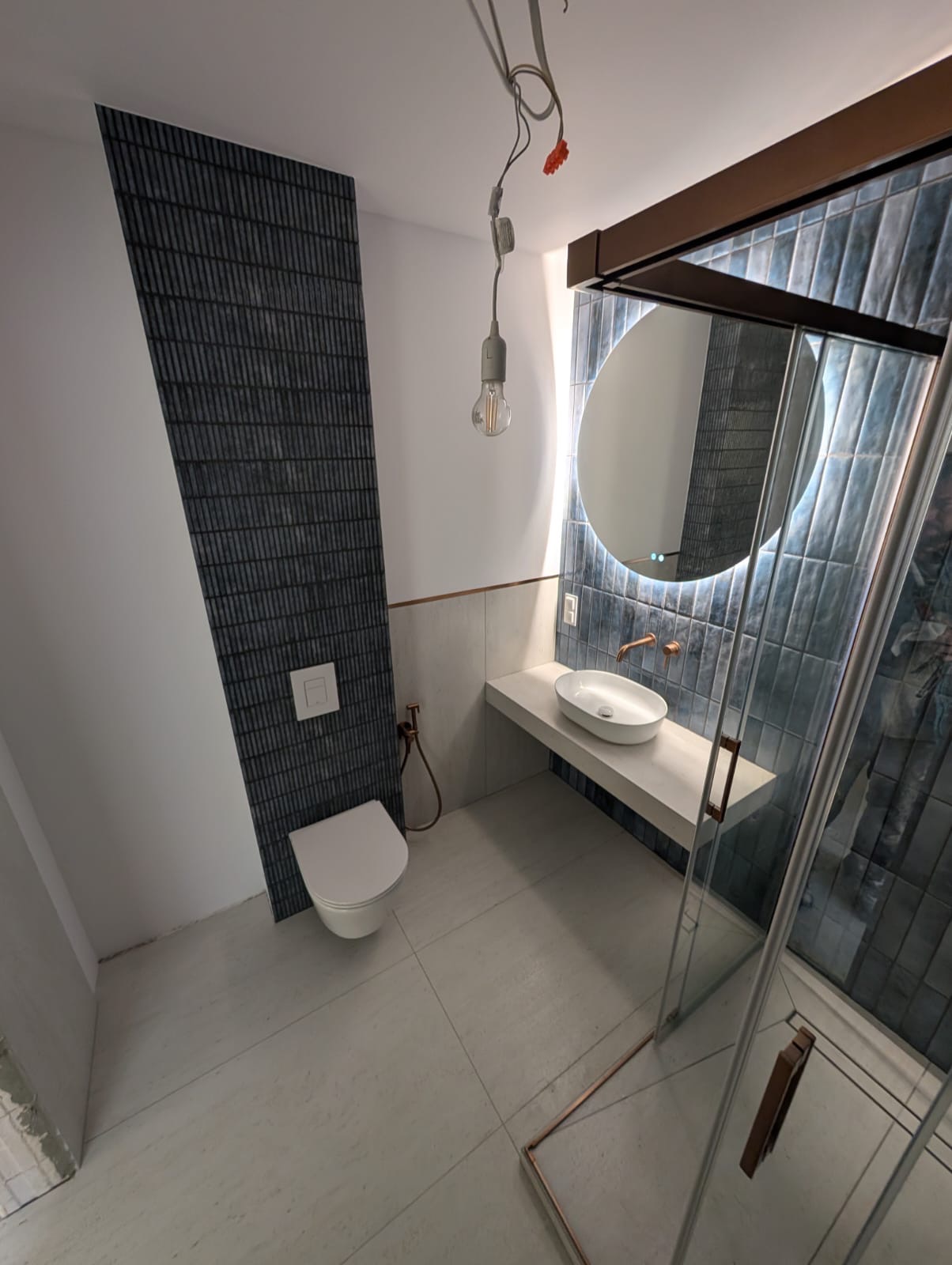 Nowoczesna łazienka z ciemnymi płytkami na ścianie, podwieszaną toaletą i umywalką na blacie. Okrągłe lustro z podświetleniem LED. Widoczna kabina prysznicowa.