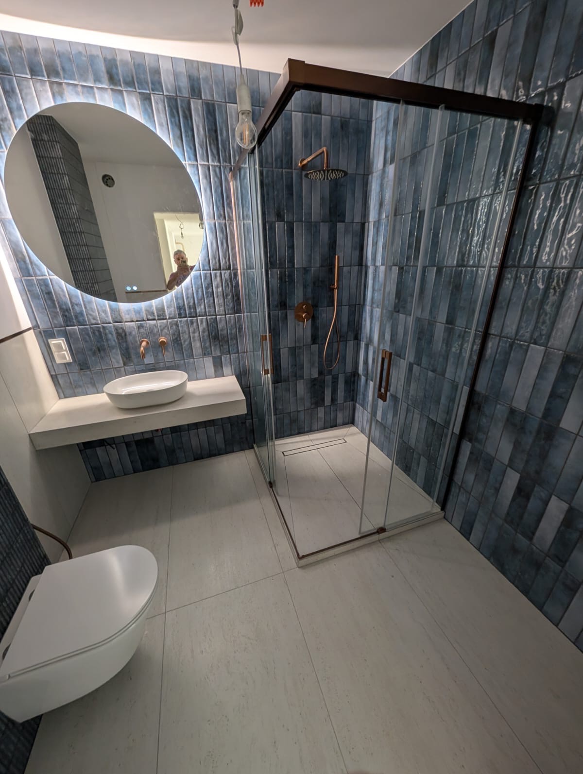 Nowoczesna łazienka z kabiną prysznicową typu walk-in, umywalką na blacie i okrągłym lustrem z podświetleniem LED. Ściany wyłożone niebieskimi, wąskimi płytkami.