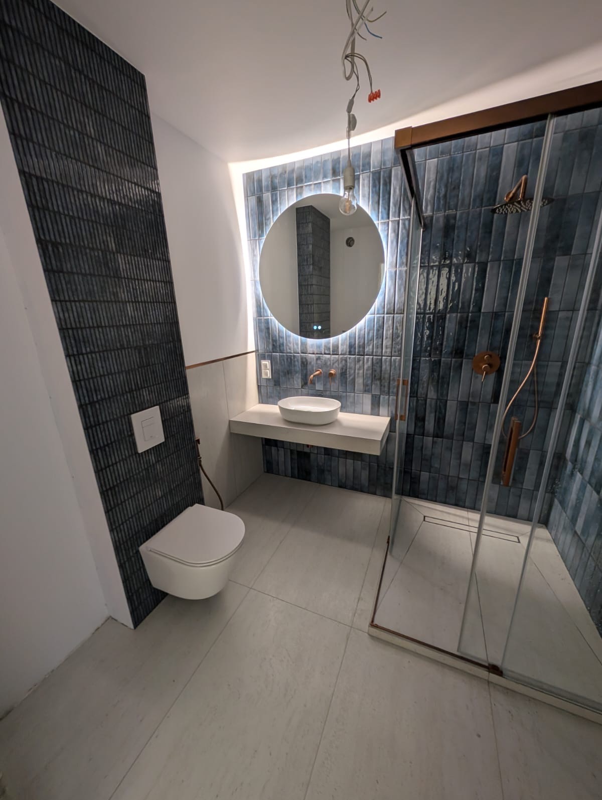 Nowoczesna łazienka z niebieskimi płytkami imitującymi cegłę, okrągłym lustrem z podświetleniem LED i kabiną prysznicową typu walk-in z miedzianymi akcentami.