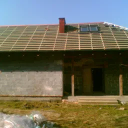 Dom w trakcie budowy dachu: widoczna drewniana konstrukcja więźby, komin z czerwonej cegły, okno dachowe i ściany z szarego bloczka betonowego. Na pierwszym planie resztki folii budowlanej.