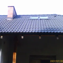 Ciemna dachówka ceramiczna na skośnym dachu z dwoma oknami dachowymi i kominem z cegły, widoczne fragmenty rynny i ściany budynku.