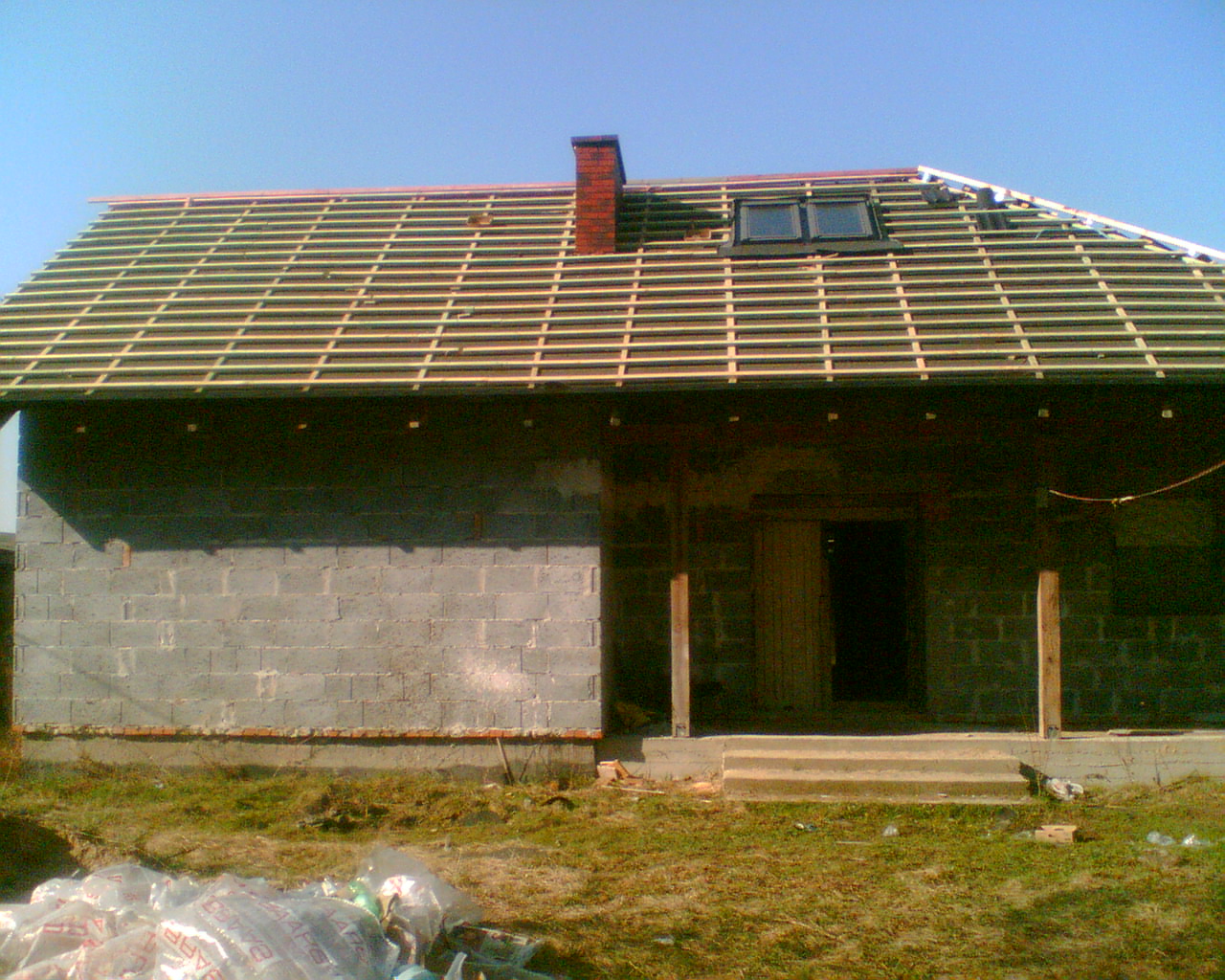 Dom w trakcie budowy dachu: widoczna drewniana konstrukcja więźby, komin z czerwonej cegły, okno dachowe i ściany z szarego bloczka betonowego. Na pierwszym planie resztki folii budowlanej.