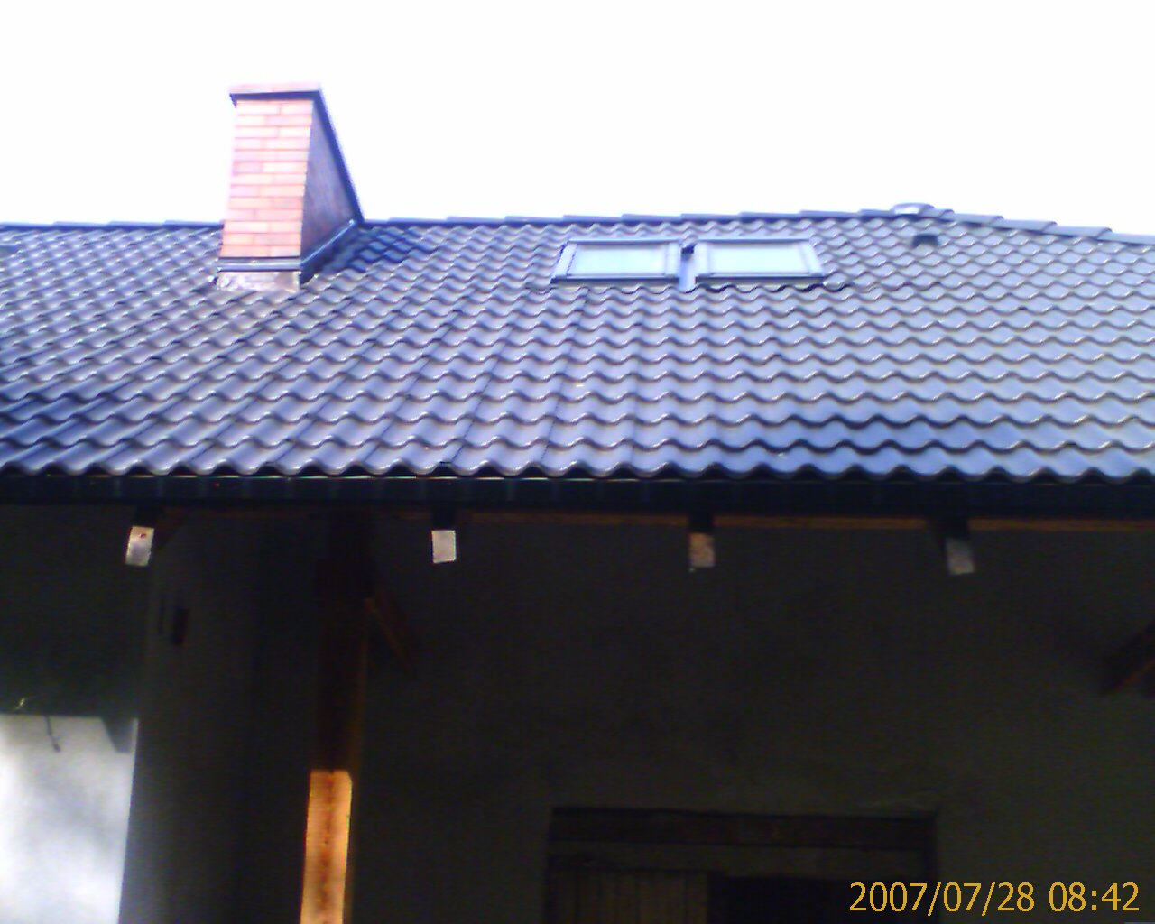 Ciemna dachówka ceramiczna na skośnym dachu z dwoma oknami dachowymi i kominem z cegły, widoczne fragmenty rynny i ściany budynku.