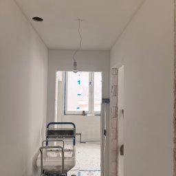 FINALITO - Wykończenia i Budownictwo Oskar Bożys - Wnętrze w trakcie malowania natryskowego na biało. Widoczny agregat malarski, okno zabezpieczone folią i surowy otwór drzwiowy. Na suficie otwór na oświetlenie.