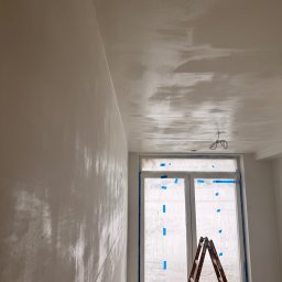 FINALITO - Wykończenia i Budownictwo Oskar Bożys - Świeżo otynkowane ściany i sufit w jasnym pokoju, zabezpieczone okno z drabiną. Widoczne ślady po gładzi szpachlowej. W tle przewody oświetleniowe.