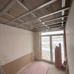 FINALITO - Wykończenia i Budownictwo Oskar Bożys - Wnętrze w trakcie prac wykończeniowych: metalowy stelaż sufitu podwieszanego, częściowo zamontowane płyty na ścianie, okno z widokiem na zimowy krajobraz.