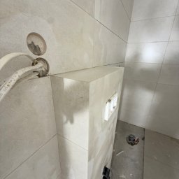 FINALITO - Wykończenia i Budownictwo Oskar Bożys - Wykończona łazienka w trakcie prac: ułożone płytki na ścianach i podłodze, widoczne rury i instalacja podtynkowa WC. Minimalistyczny design w jasnych odcieniach.