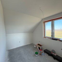 FINALITO - Wykończenia i Budownictwo Oskar Bożys - Pokój na poddaszu po malowaniu na biało, widoczne ślady farby na podłodze, okno z widokiem na pole, puszki z farbą i narzędzia malarskie.