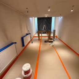 FINALITO - Wykończenia i Budownictwo Oskar Bożys - Przygotowania do malowania podłogi: zabezpieczone listwy i grzejniki taśmą, widoczne puszki z farbą i drabina w głębi pomieszczenia. Oświetlenie sufitowe.