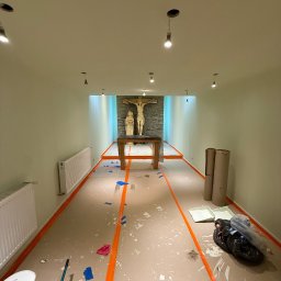 FINALITO - Wykończenia i Budownictwo Oskar Bożys - Wnętrze kaplicy w trakcie malowania: zabezpieczona podłoga taśmą, widoczne narzędzia malarskie, ściany w jasnym odcieniu, w tle krucyfiks i figury.