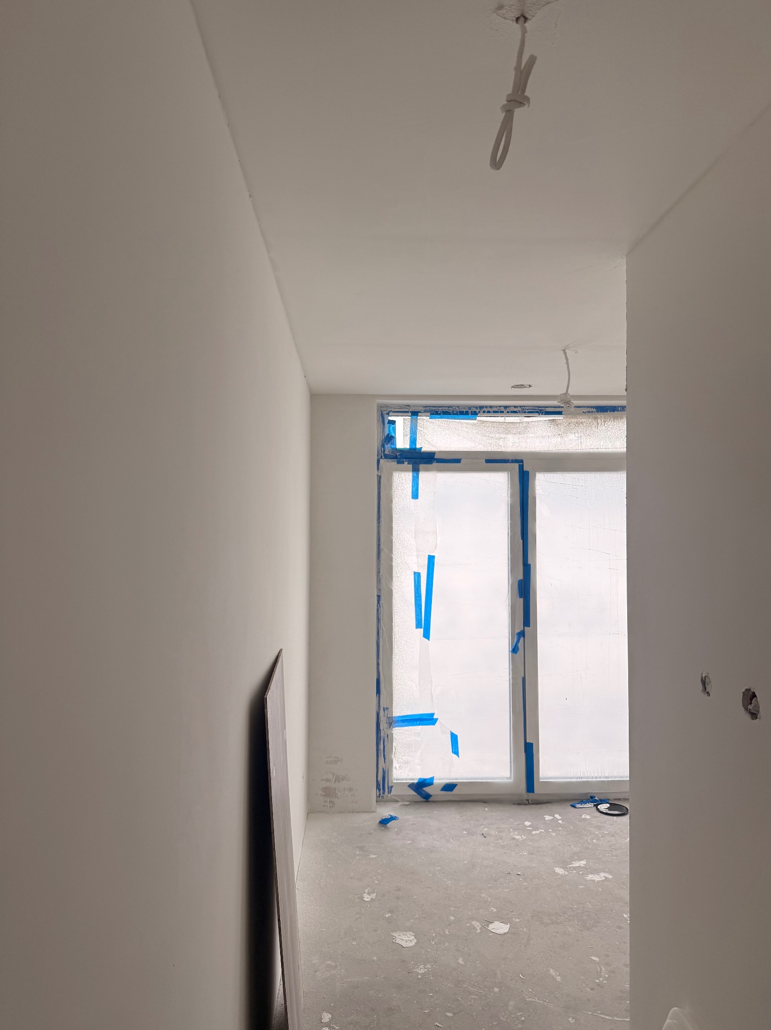 Wnętrze w trakcie prac wykończeniowych: zabezpieczone okno folią i taśmą, świeżo pomalowane ściany, widoczne kable i płyta karton-gips oparta o ścianę.