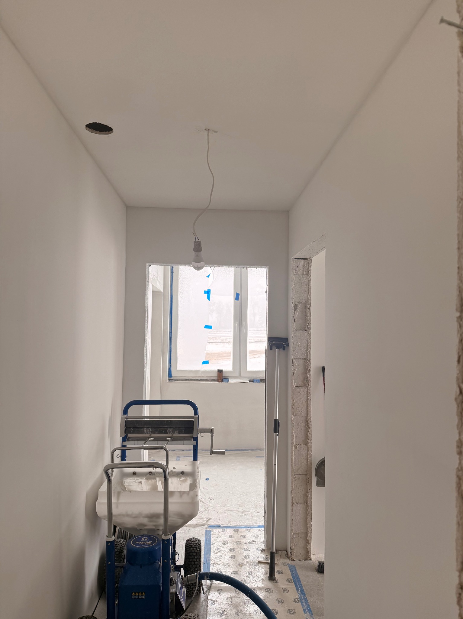 Wnętrze w trakcie malowania natryskowego na biało. Widoczny agregat malarski, okno zabezpieczone folią i surowy otwór drzwiowy. Na suficie otwór na oświetlenie.