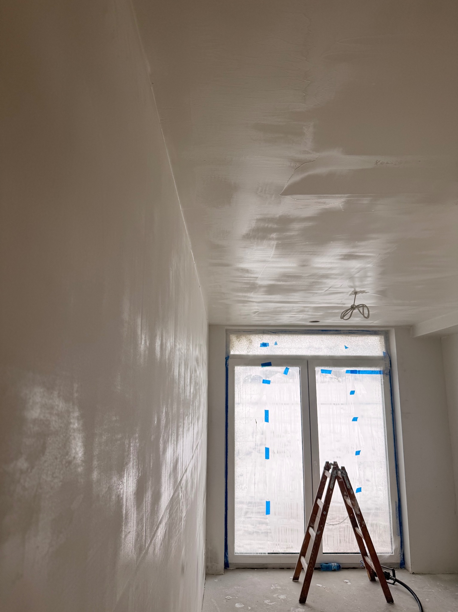 Świeżo otynkowane ściany i sufit w jasnym pokoju, zabezpieczone okno z drabiną. Widoczne ślady po gładzi szpachlowej. W tle przewody oświetleniowe.