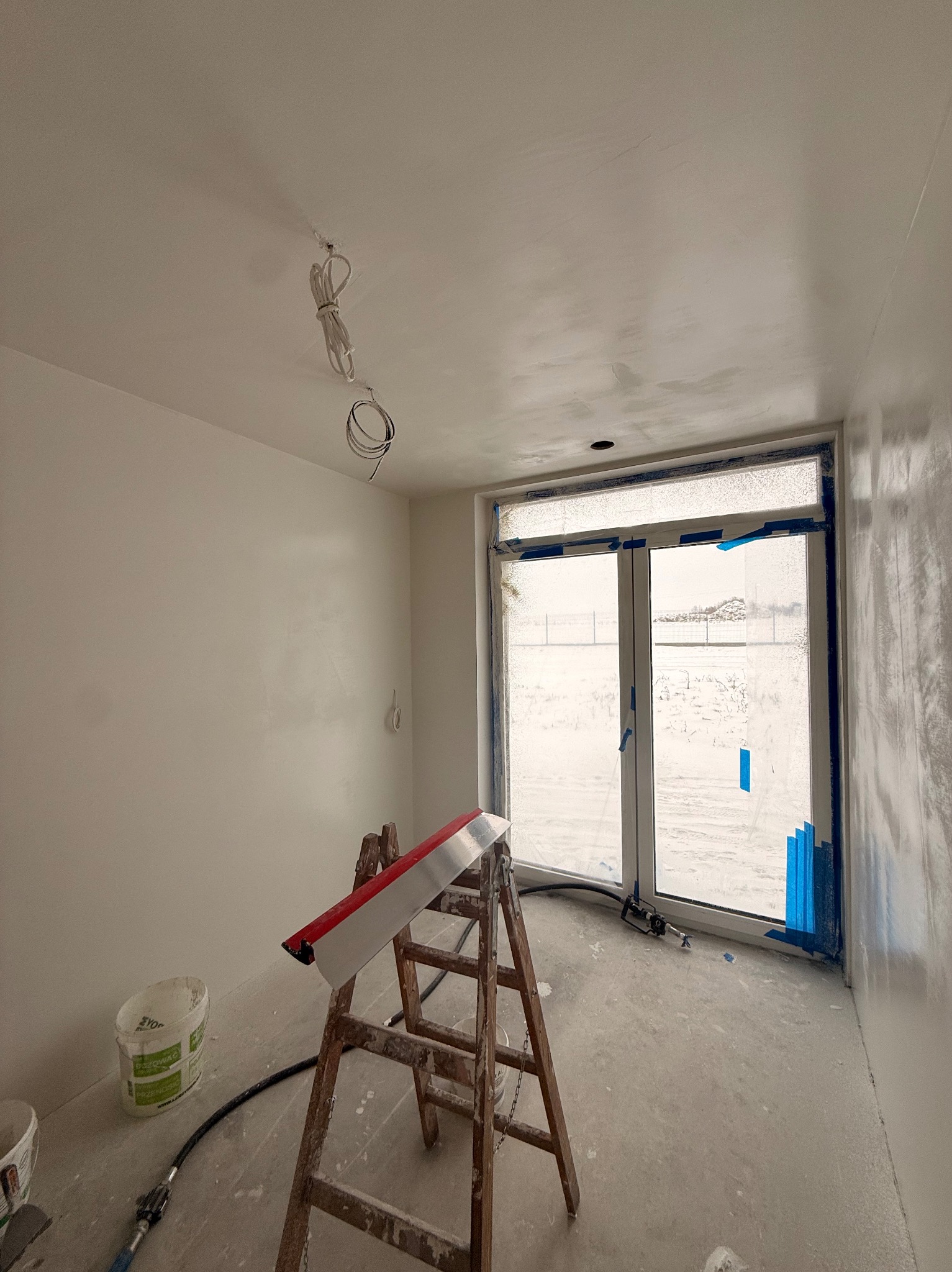 Pokój po gładziowaniu ścian, z drabiną, narzędziami i oknem zabezpieczonym taśmą malarską. Widoczne niedoskonałości gładzi przed szlifowaniem.