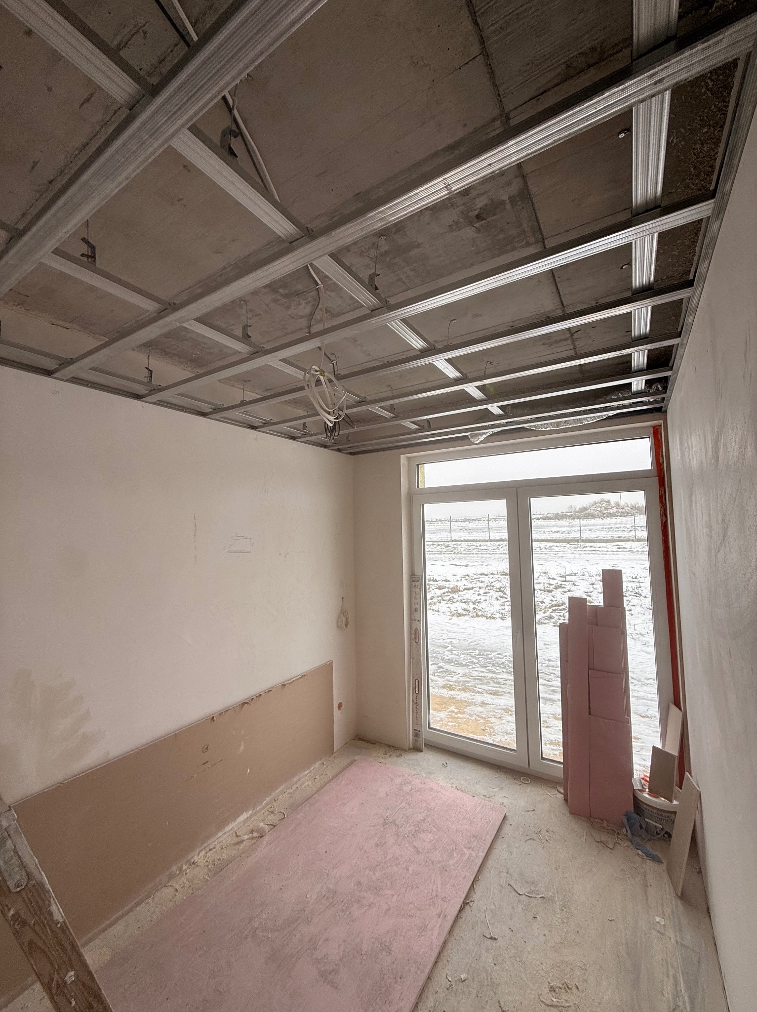 Wnętrze w trakcie prac wykończeniowych: metalowy stelaż sufitu podwieszanego, częściowo zamontowane płyty na ścianie, okno z widokiem na zimowy krajobraz.