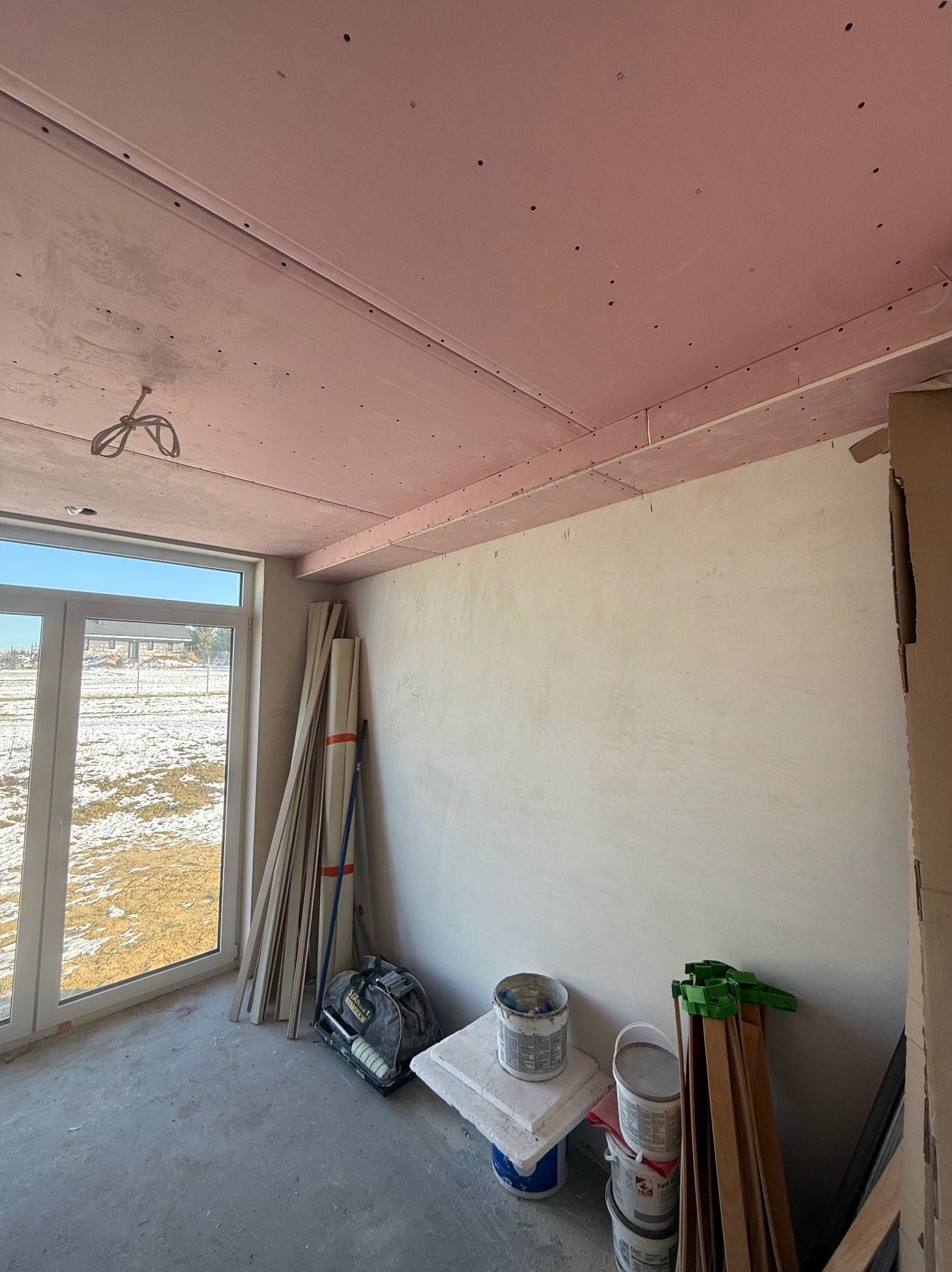 Wnętrze w trakcie prac wykończeniowych: sufit z płyt karton-gips w kolorze różowym, ściany przygotowane do malowania, widoczne narzędzia i materiały budowlane.