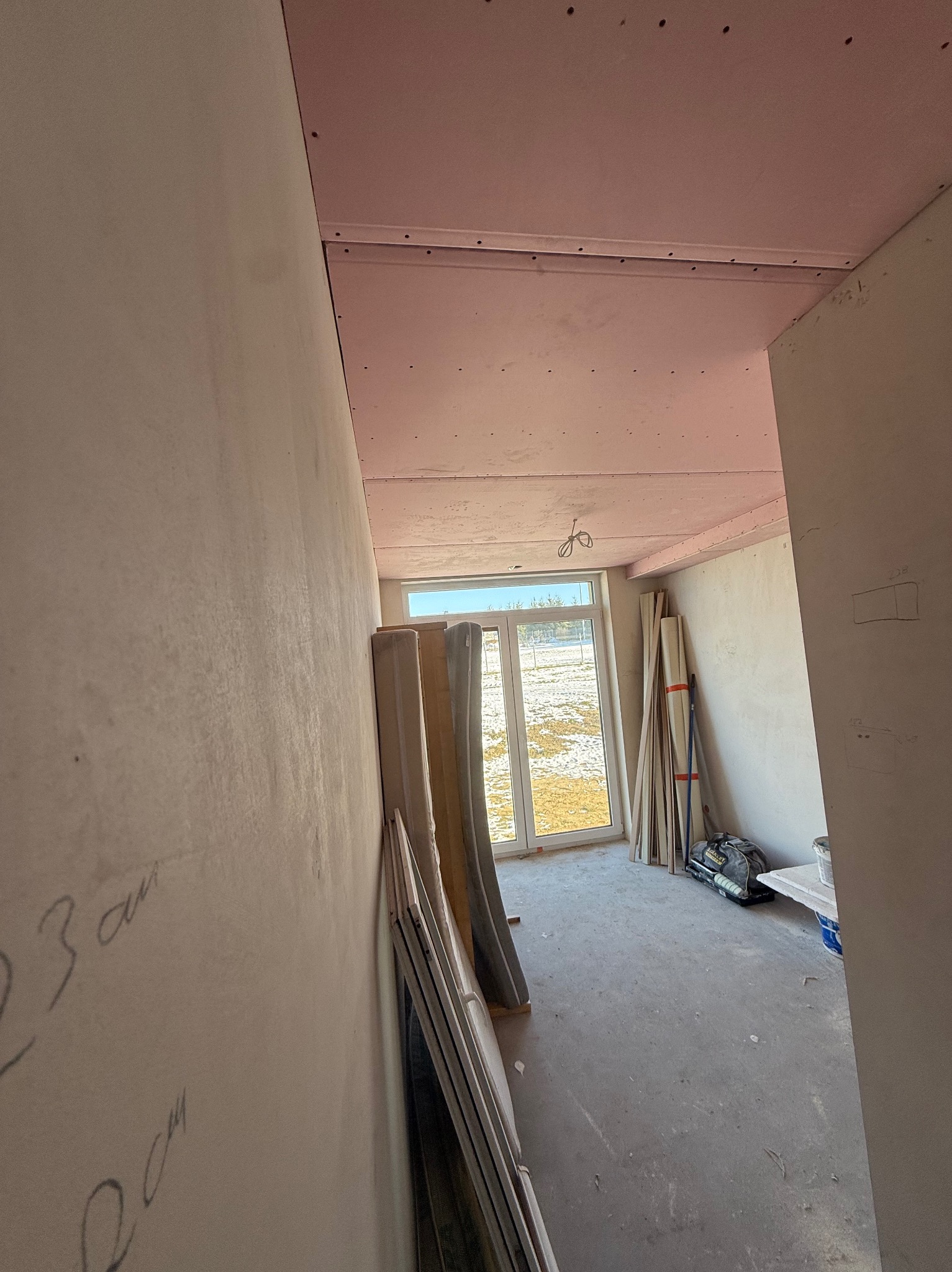 Wnętrze w trakcie prac wykończeniowych: sufit z płyt karton-gips w kolorze różowym, ściany przygotowane do malowania, okno z widokiem na zewnątrz, materiały budowlane oparte o ścianę.