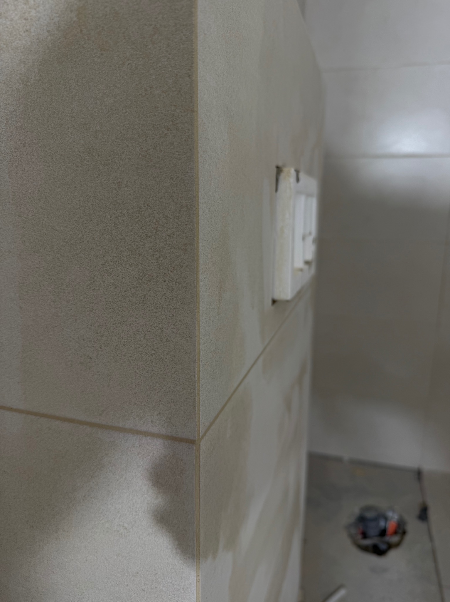 Beżowe płytki ścienne z fugami, narożnik i gniazdka elektryczne w łazience w trakcie remontu. Widoczny fragment podłogi z odpływem.