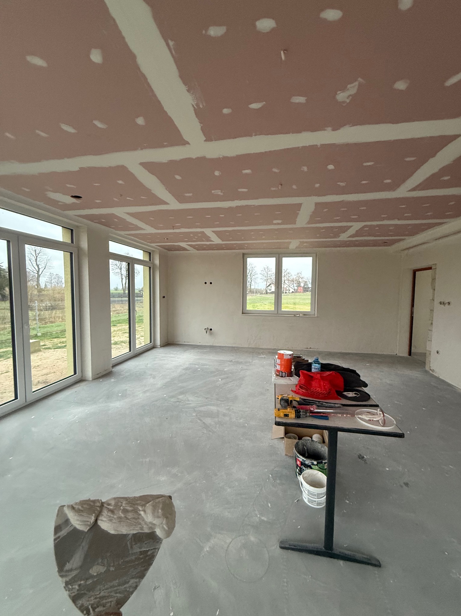 Wnętrze w trakcie prac wykończeniowych: sufit z płyt karton-gips, szpachlowane łączenia, narzędzia na stole. Widok na okna i krajobraz za nimi.