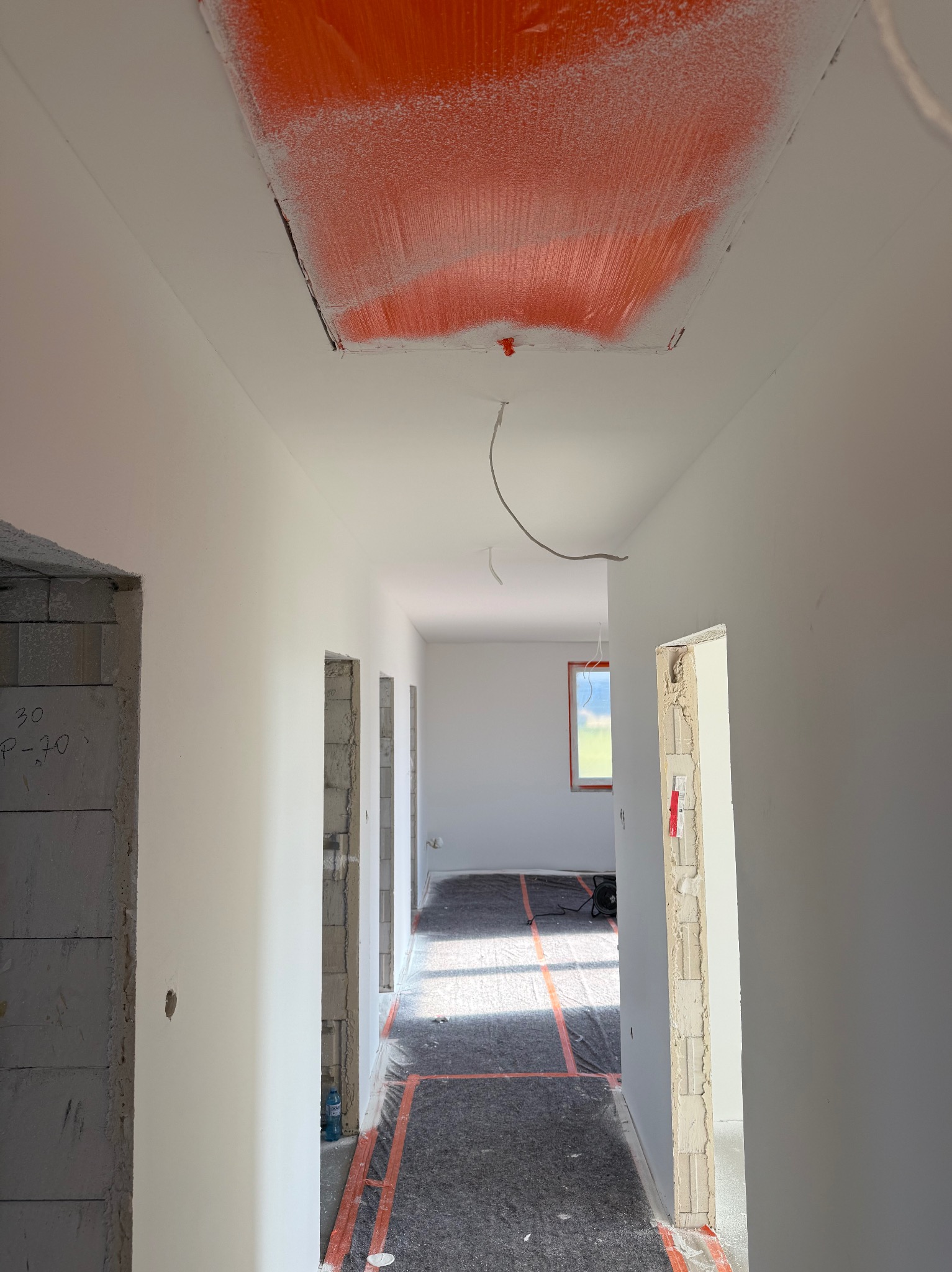 Wnętrze w trakcie prac wykończeniowych: białe ściany, przygotowana podłoga z folią w kratę, sufit z pomarańczowym elementem. Widoczne surowe ościeżnice.