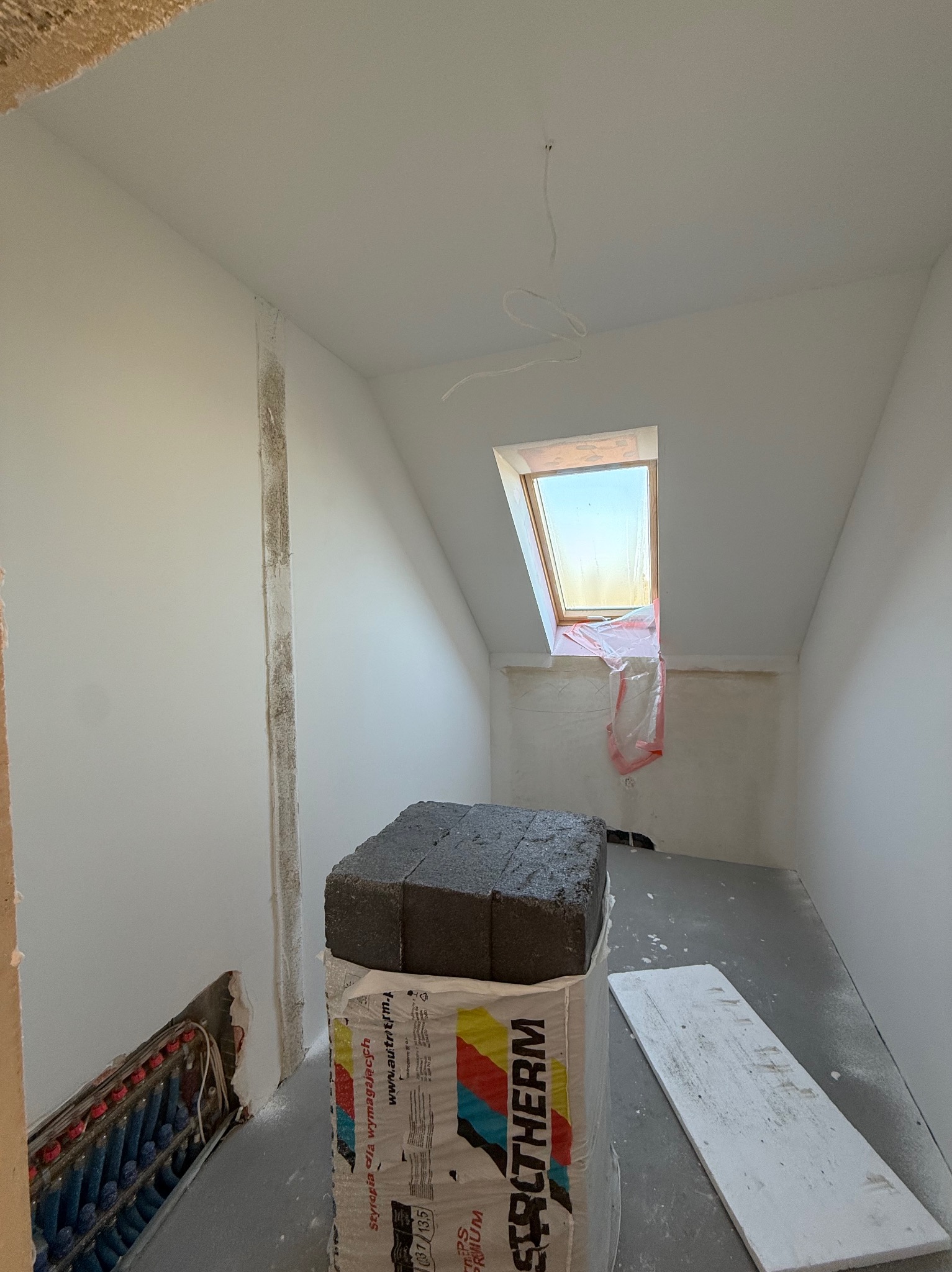 Poddasze w trakcie prac wykończeniowych: ocieplenie, instalacja ogrzewania podłogowego, okno dachowe zabezpieczone folią. Widoczne materiały izolacyjne.