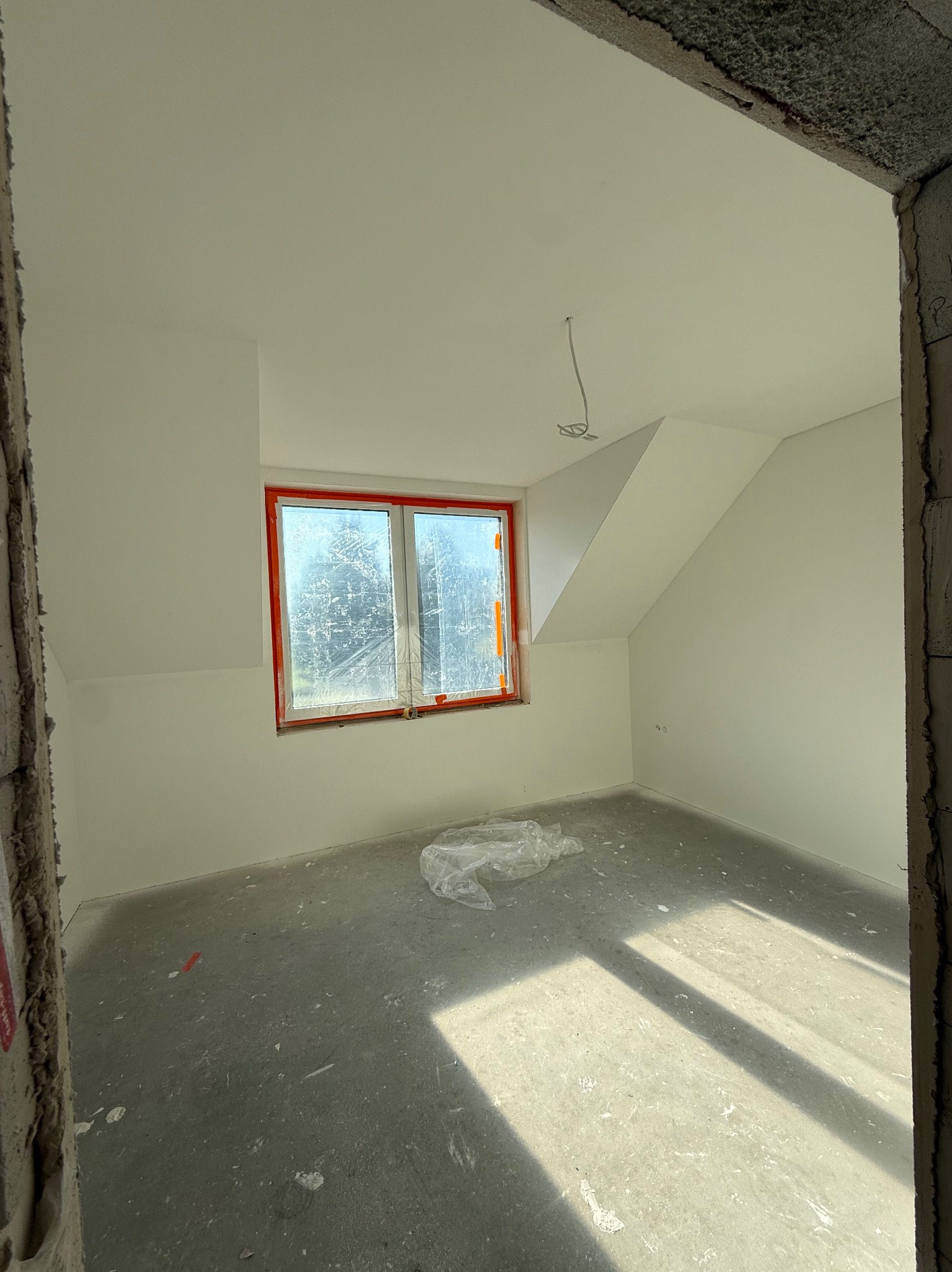 Pokój na poddaszu po malowaniu ścian na biało, z oknem zabezpieczonym folią i surową podłogą. Widoczne ślady po pracach wykończeniowych.