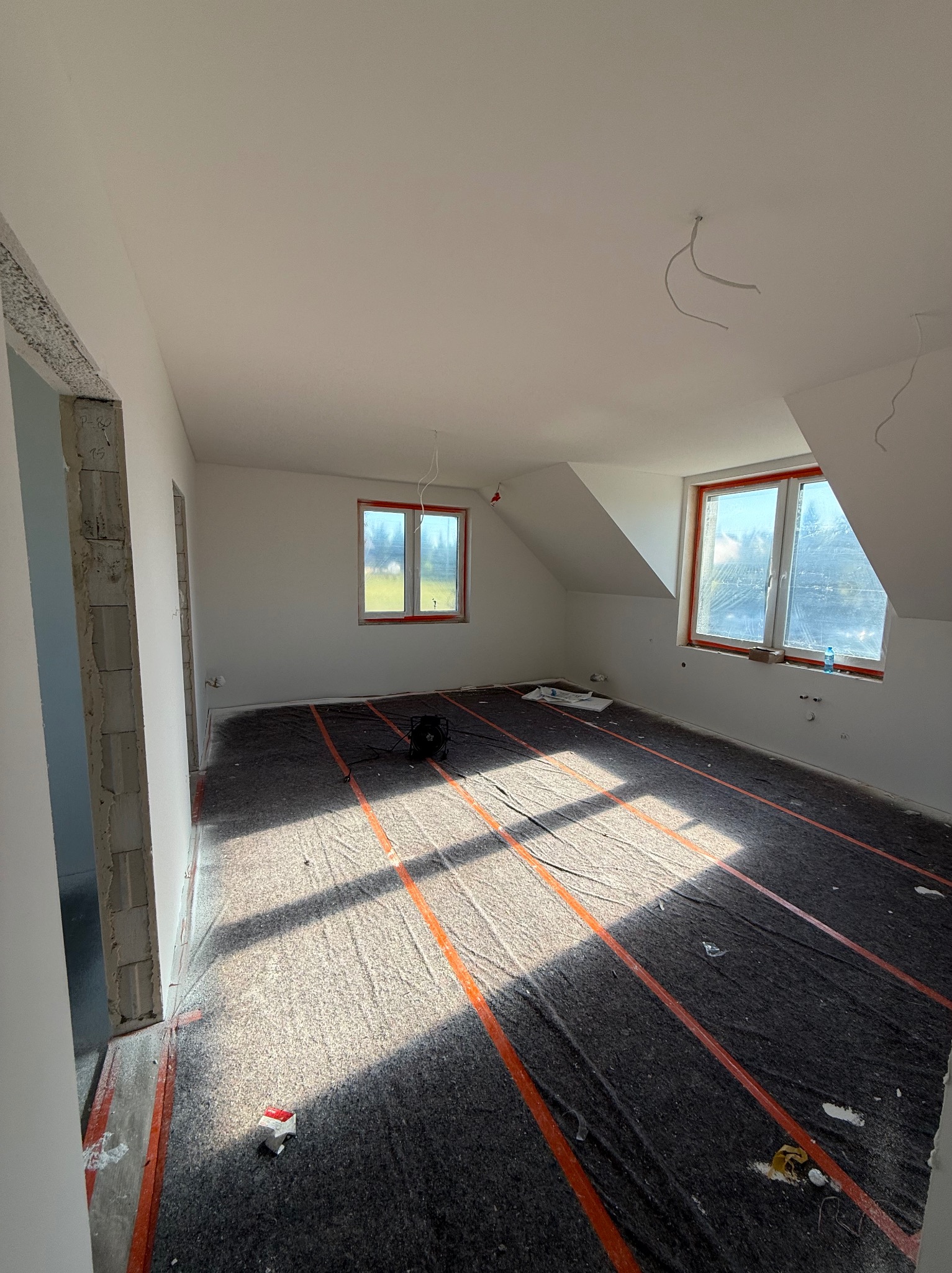 Pokój na poddaszu w trakcie prac wykończeniowych. Widoczne przygotowanie pod wylewkę, okna z folią ochronną i surowe ściany. W tle widok na zewnątrz.