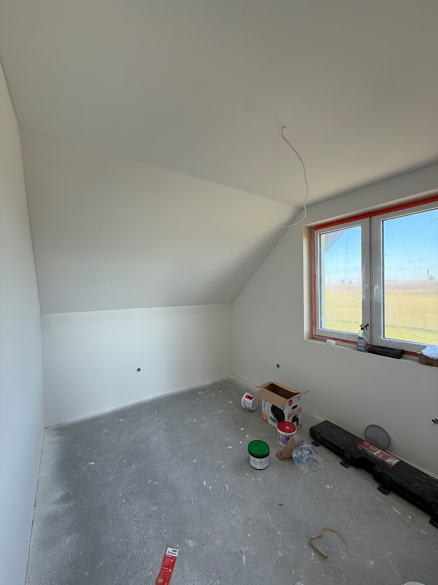 Pokój na poddaszu po malowaniu na biało, widoczne ślady farby na podłodze, okno z widokiem na pole, puszki z farbą i narzędzia malarskie.