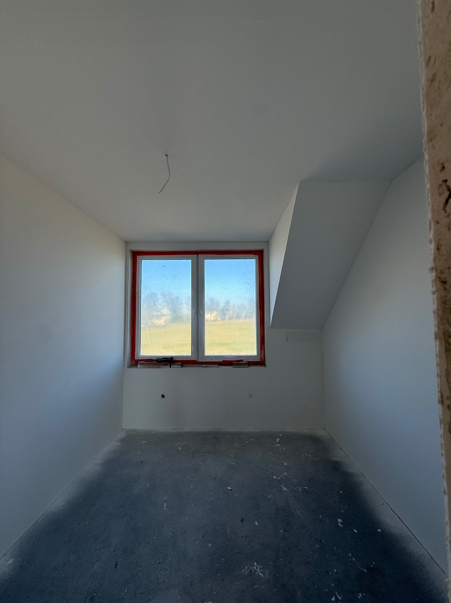Wnętrze pokoju na poddaszu po malowaniu ścian na biało. Okno z czerwoną taśmą zabezpieczającą. Szara podłoga. Widoczny fragment surowej ściany.
