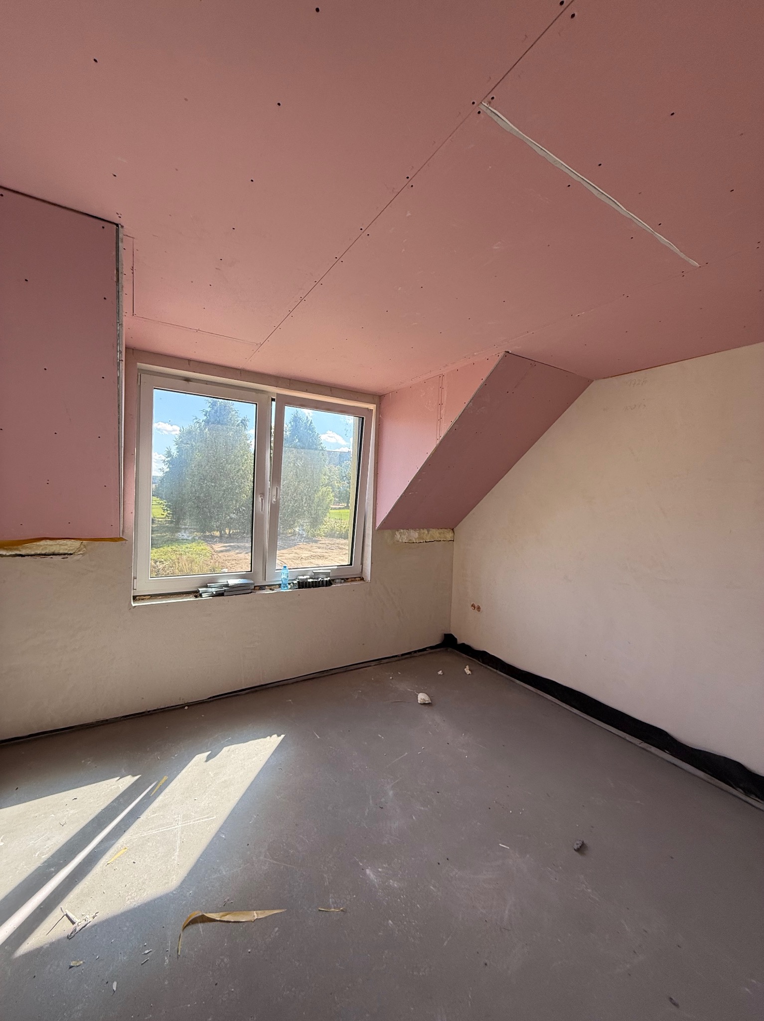 Pokój na poddaszu z oknem, ściany częściowo wykończone gładzią, sufit i fragmenty ścian pokryte różową płytą karton-gips. Widoczne narzędzia przy oknie.
