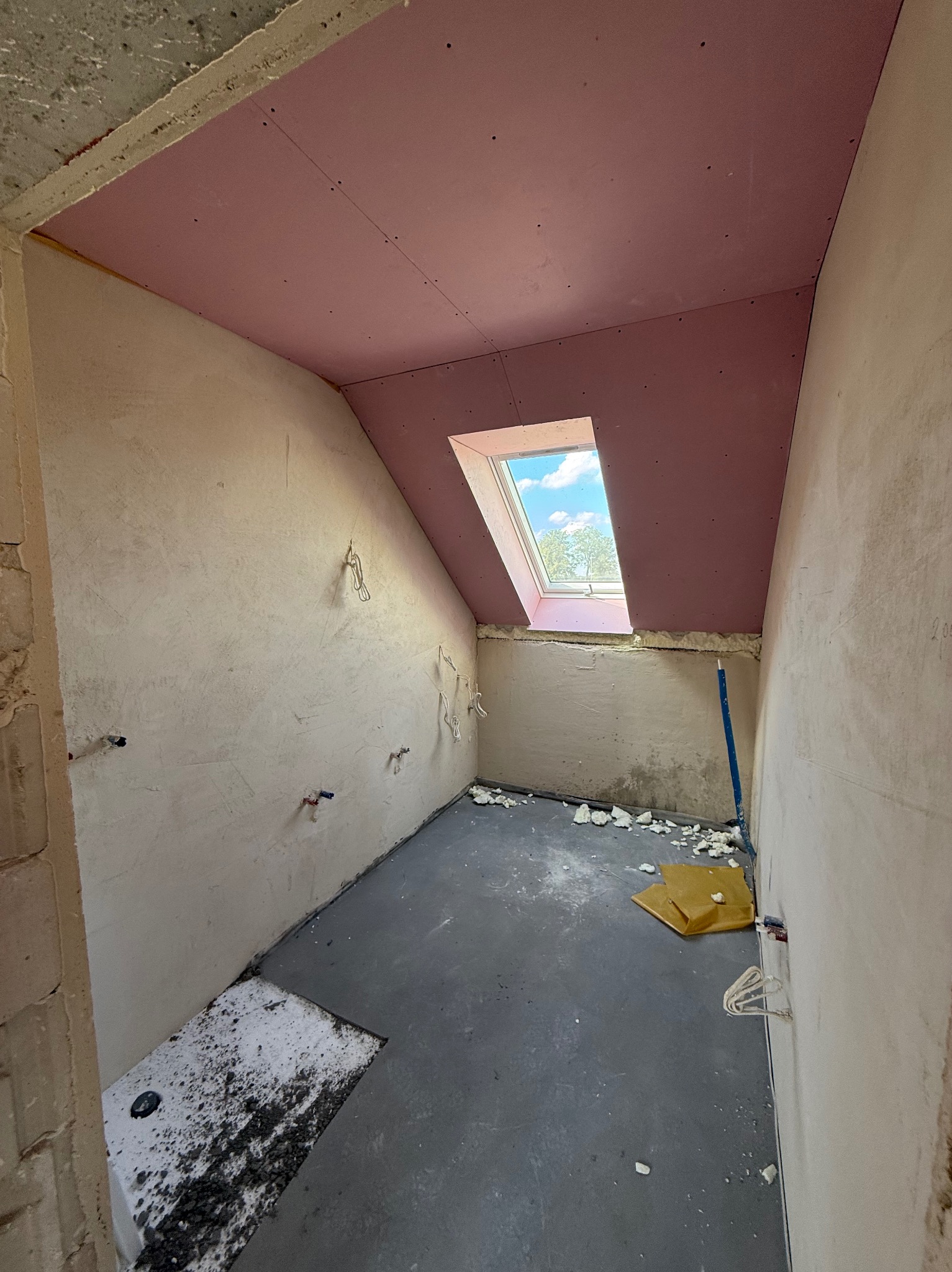 Poddasze w trakcie wykańczania: różowe płyty karton-gips na skosach sufitu, surowe ściany, okno dachowe z widokiem na niebo. Etap prac wykończeniowych.