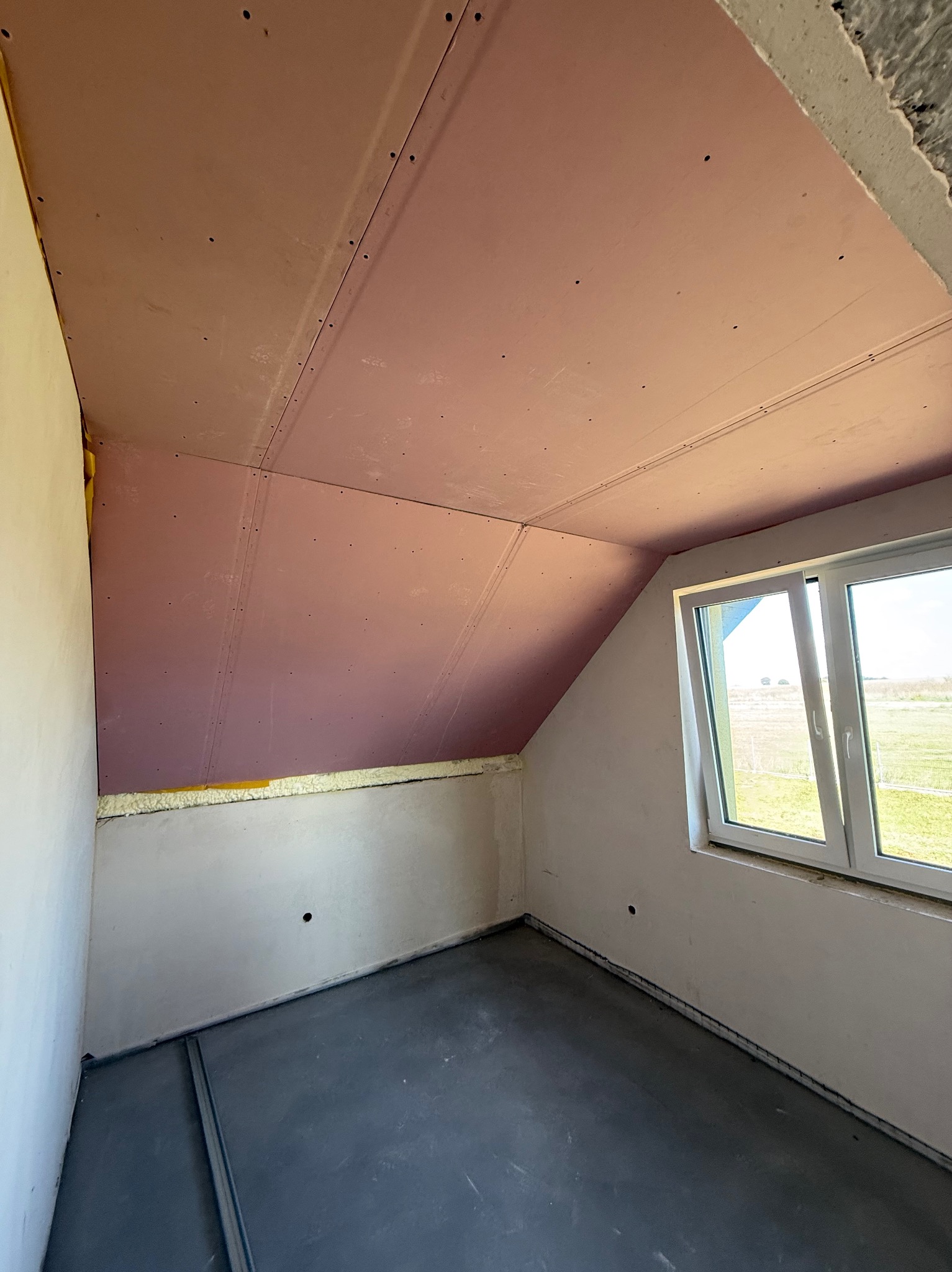 Poddasze z oknem po montażu płyt karton-gips na skosach dachu. Widoczne łączenia płyt i szpary do wypełnienia. Na podłodze wylewka. Okno z widokiem na zewnątrz.