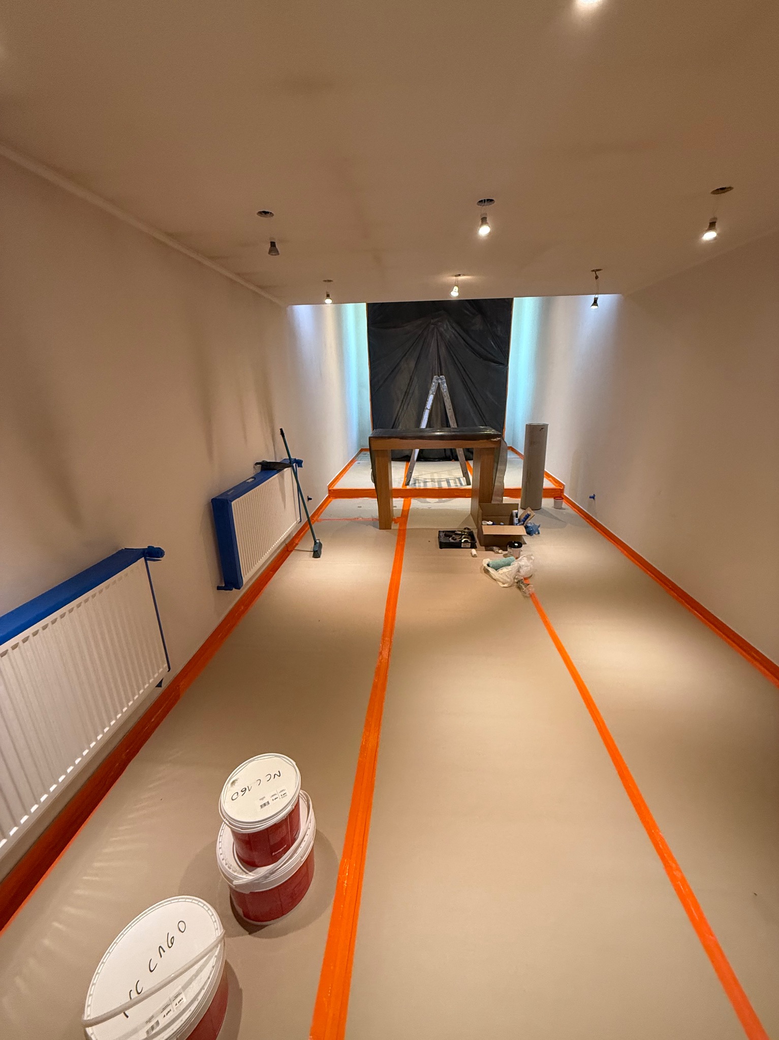 Przygotowania do malowania podłogi: zabezpieczone listwy i grzejniki taśmą, widoczne puszki z farbą i drabina w głębi pomieszczenia. Oświetlenie sufitowe.