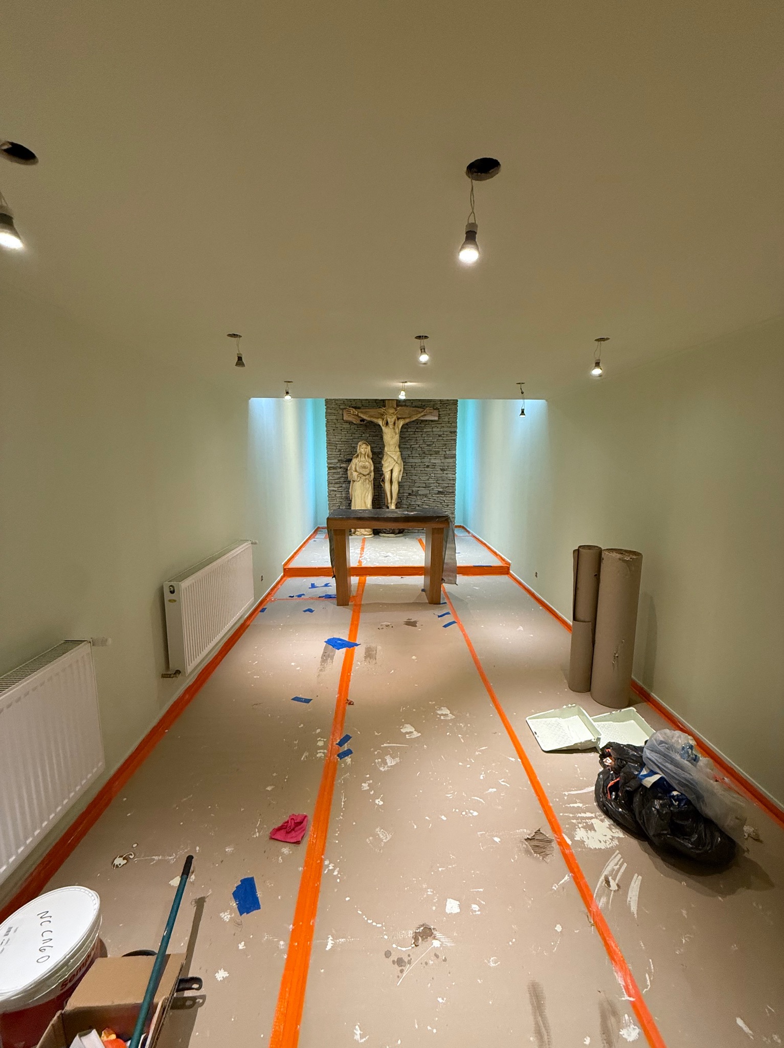 Wnętrze kaplicy w trakcie malowania: zabezpieczona podłoga taśmą, widoczne narzędzia malarskie, ściany w jasnym odcieniu, w tle krucyfiks i figury.