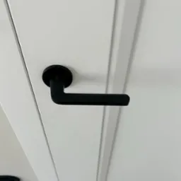 Białe drzwi wewnętrzne z czarną, minimalistyczną klamką. Widoczne frezowane panele drzwiowe i fragment sufitu. Ujęcie z dołu, podkreślające nowoczesny design.