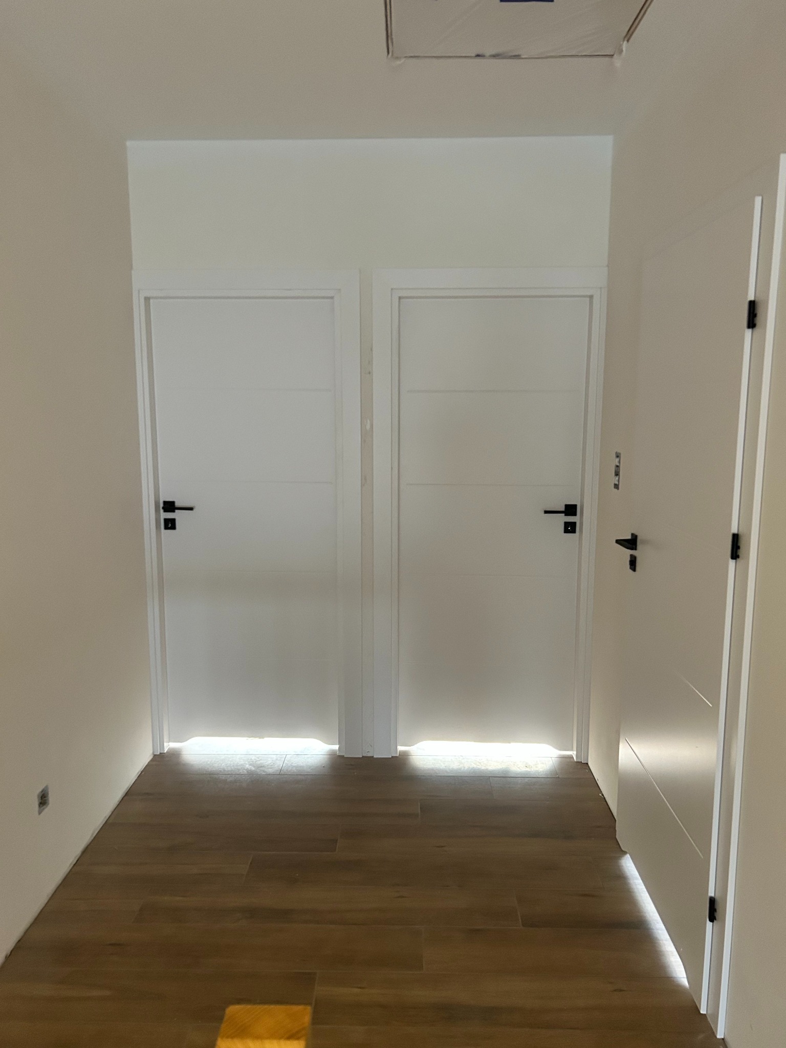Korytarz z trzema białymi drzwiami i drewnopodobną podłogą. Dwie pary drzwi są umieszczone obok siebie, trzecie drzwi po prawej stronie.