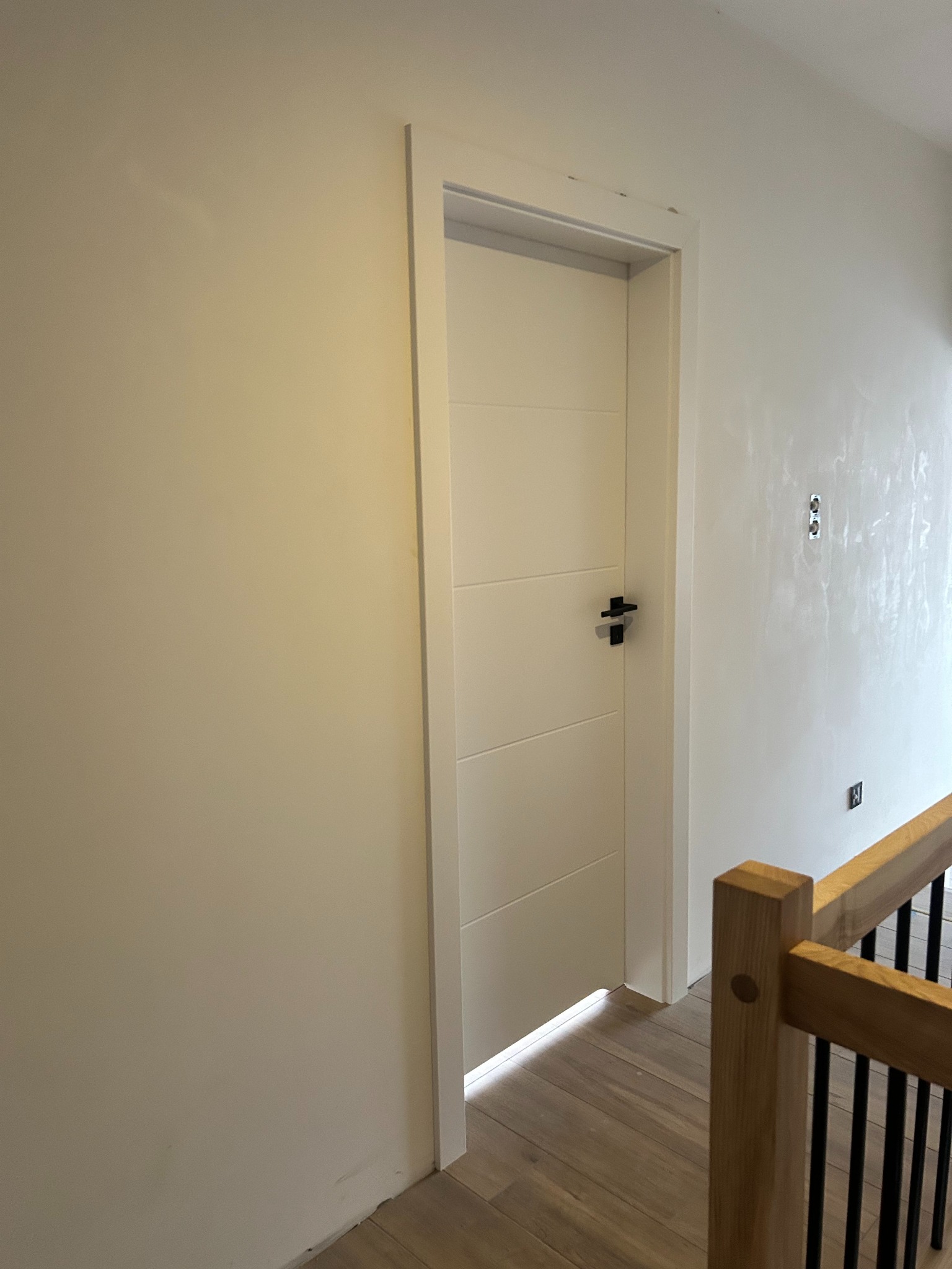 Białe, panelowe drzwi wewnętrzne z czarną klamką, zamontowane w jasnym pomieszczeniu z drewnianą podłogą i elementami balustrady. Minimalistyczny design.