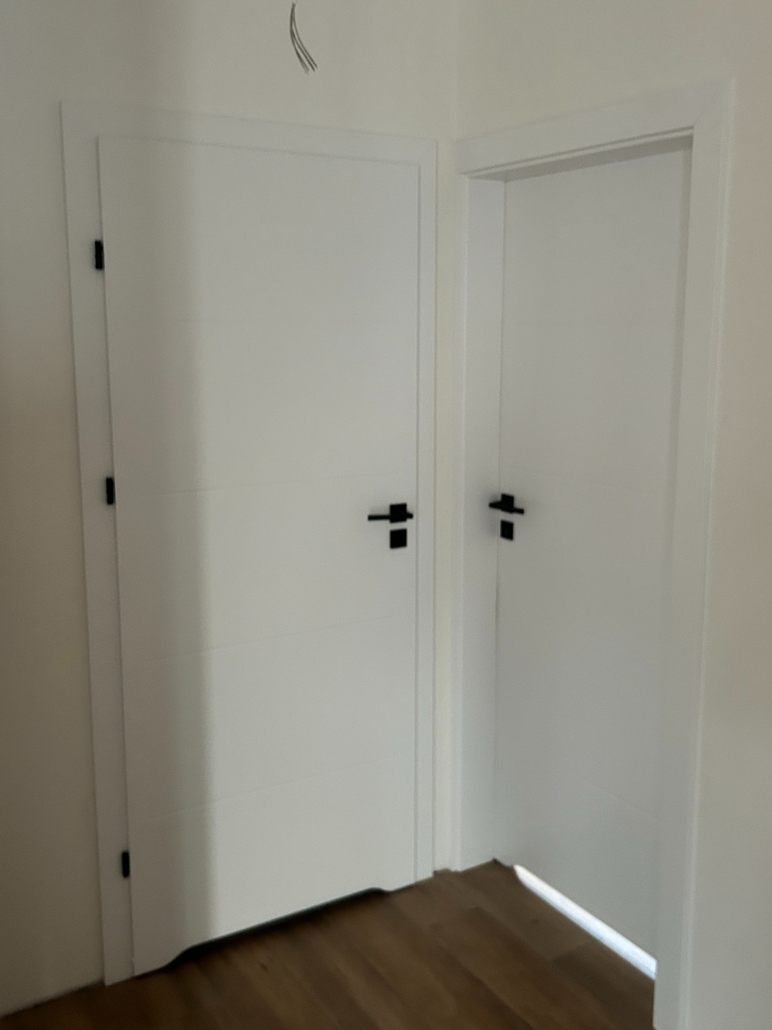 Dwa białe, matowe drzwi wewnętrzne z czarnymi klamkami na tle jasnej ściany i drewnianej podłogi, widoczne kable wystające z sufitu.