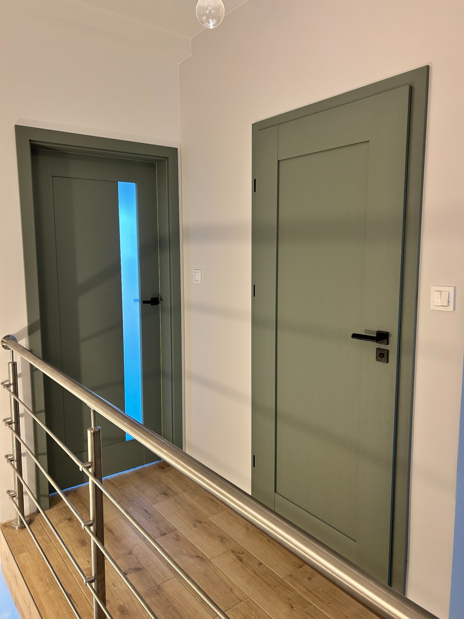 Dwa odcienie zieleni drzwi wewnętrznych z czarnymi klamkami na tle jasnych ścian i drewnopodobnej podłogi, widok zza metalowej barierki, subtelne oświetlenie.