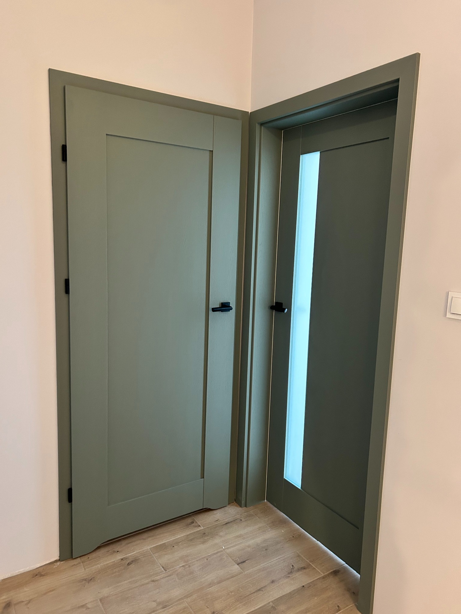 Dwa odcienie zieleni: pełne drzwi i drzwi z szybą, z czarnymi klamkami, w jasnym wnętrzu. Widoczne drewniane panele podłogowe i fragment ściany z włącznikiem.