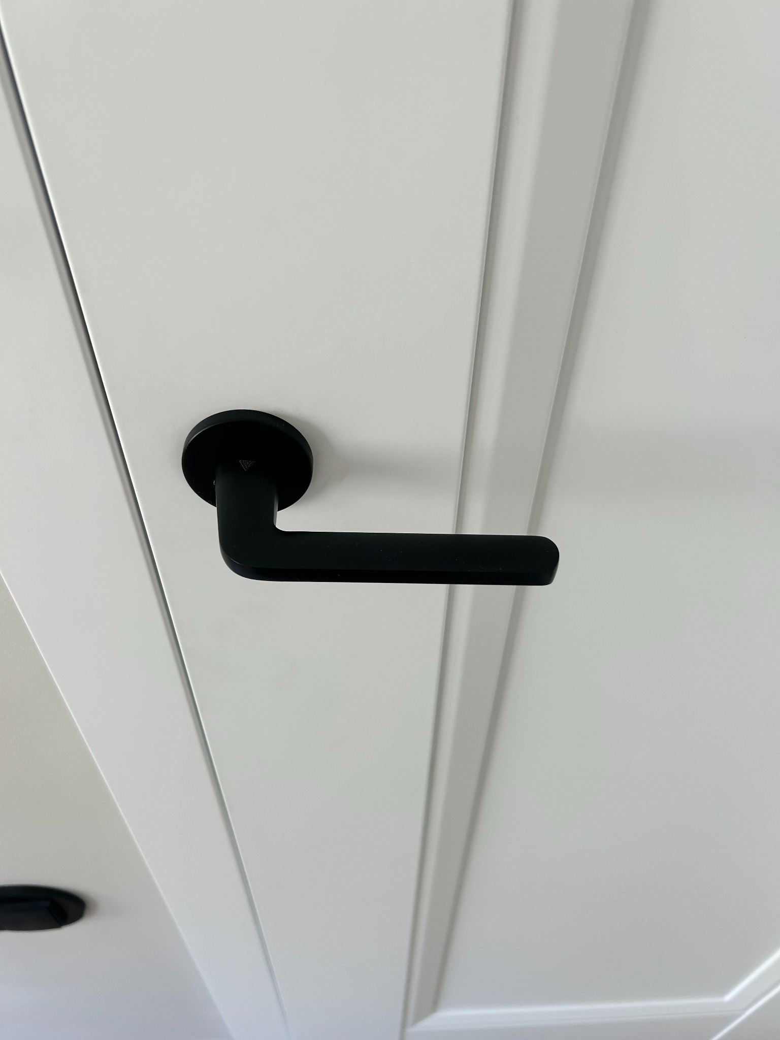 Białe drzwi wewnętrzne z czarną, minimalistyczną klamką. Widoczne frezowane panele drzwiowe i fragment sufitu. Ujęcie z dołu, podkreślające nowoczesny design.