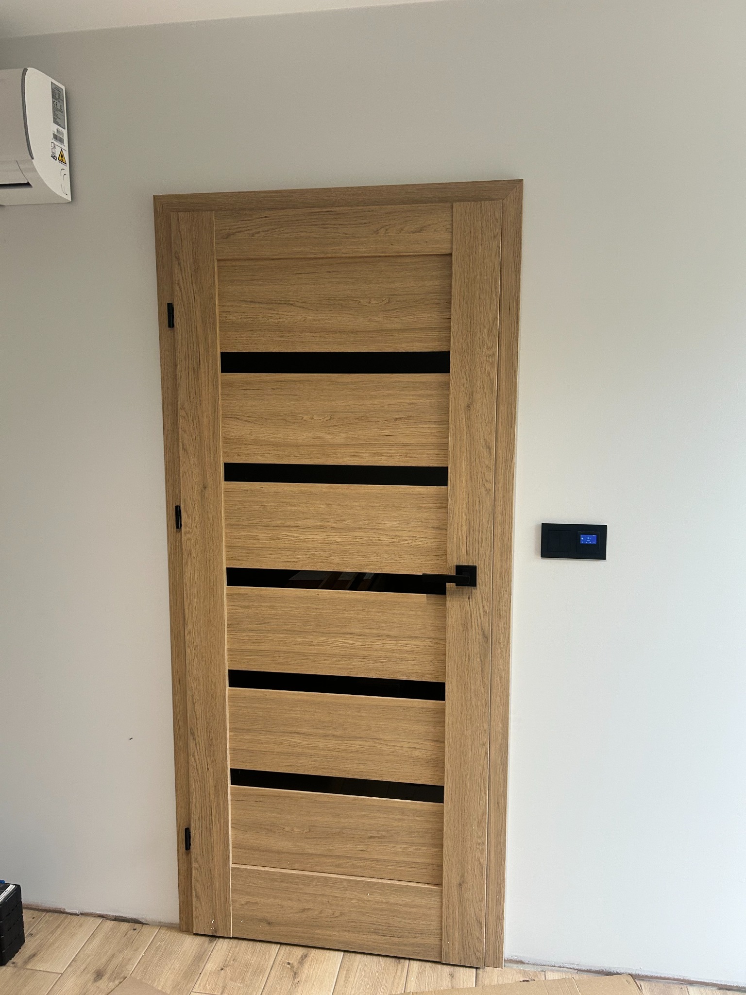 Zamknięte, dębowe drzwi wewnętrzne z czarnymi, poziomymi wstawkami, zamontowane w białej ścianie, z widocznym panelem sterowania klimatyzacją i fragmentem podłogi.
