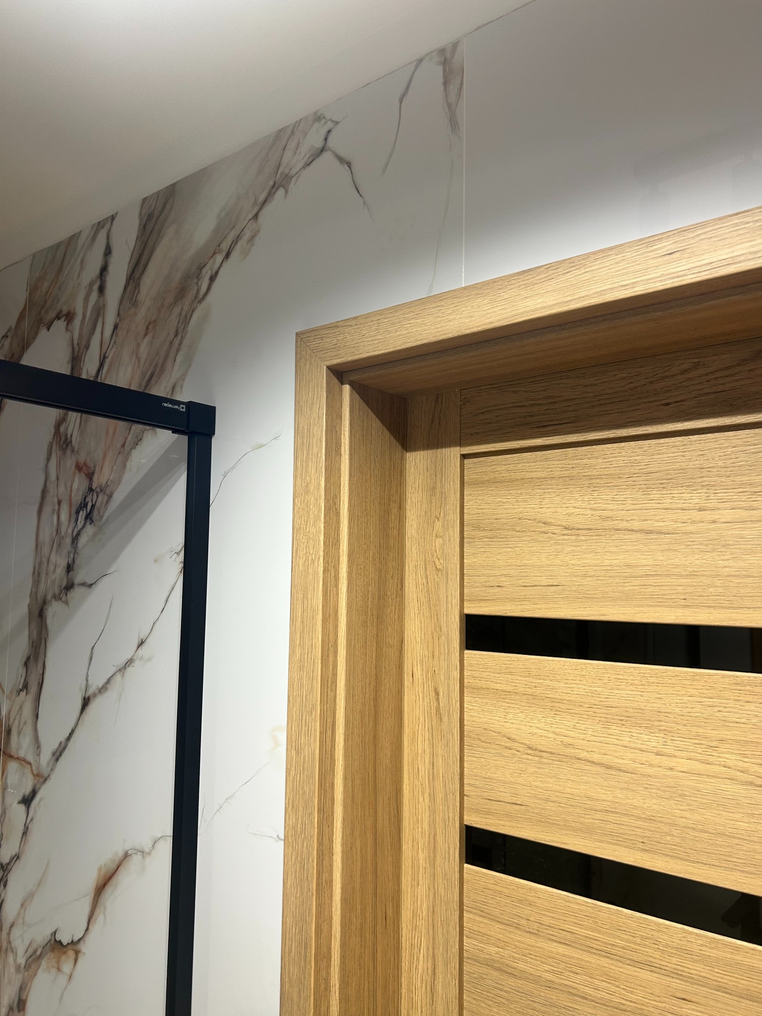 Fragment wnętrza: drzwi wewnętrzne z drewnianą okleiną i czarnymi akcentami, na tle ściany z marmurowym wzorem i fragmentem czarnej ramy kabiny prysznicowej.