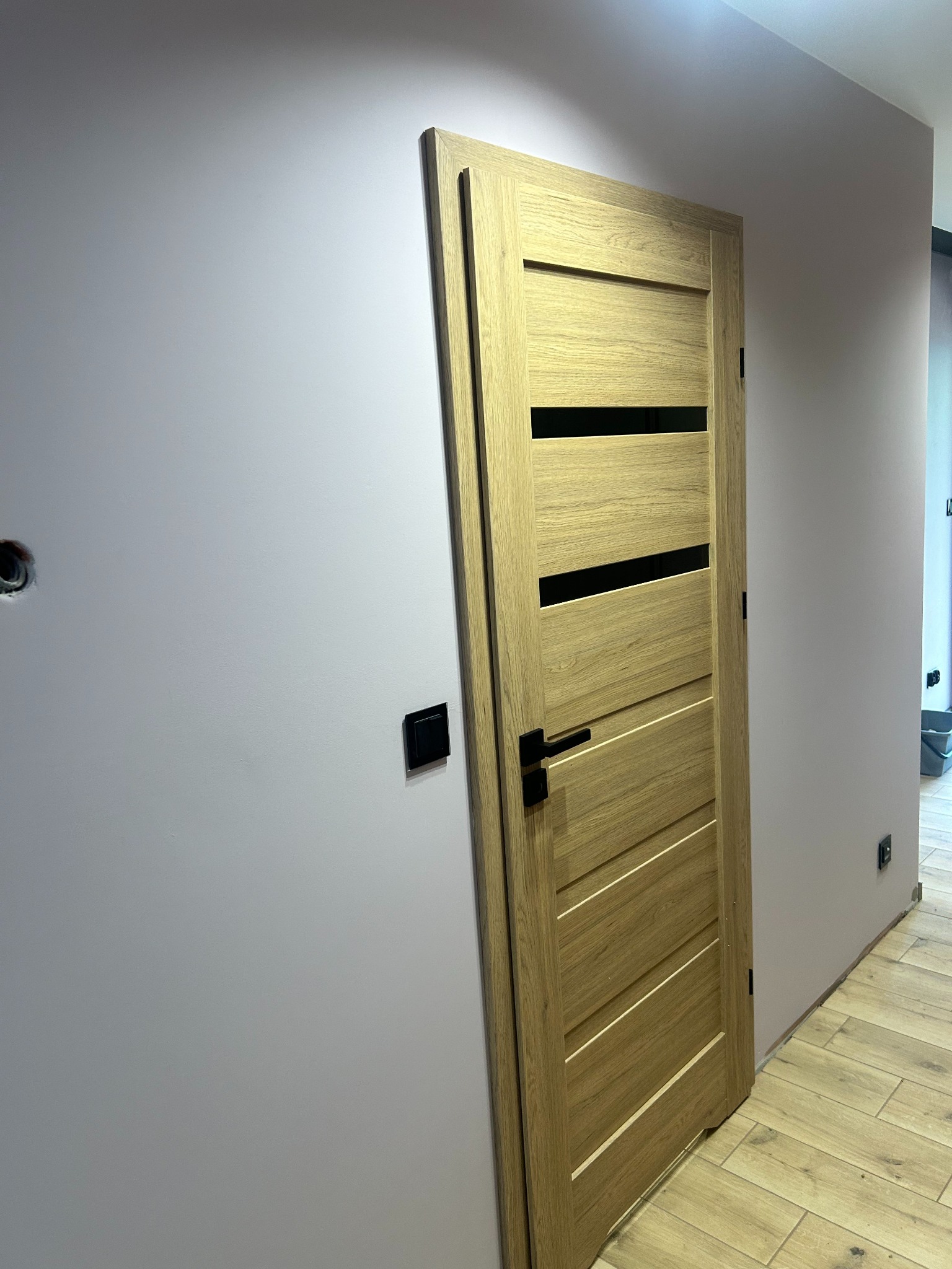 Zamontowane drzwi wewnętrzne z drewna o jasnym odcieniu, z czarnymi, poziomymi akcentami, czarną klamką na tle ściany w kolorze pudrowego różu i drewnianej podłogi.