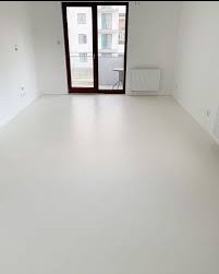 Biała, gładka posadzka w jasnym wnętrzu z oknem balkonowym i grzejnikiem. Minimalistyczny design, świeżo wykończone pomieszczenie, widok na budynek zza balkonu.