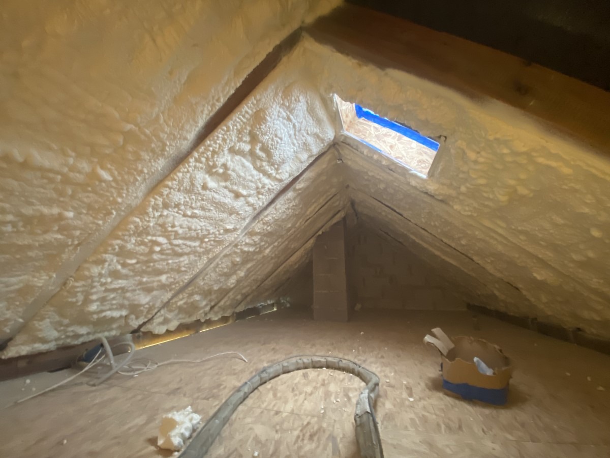 Poddasze po aplikacji pianki PUR, widoczne nierównomierne pokrycie izolacją, fragment okna dachowego z niebieską taśmą. Na podłodze leżą resztki materiałów.