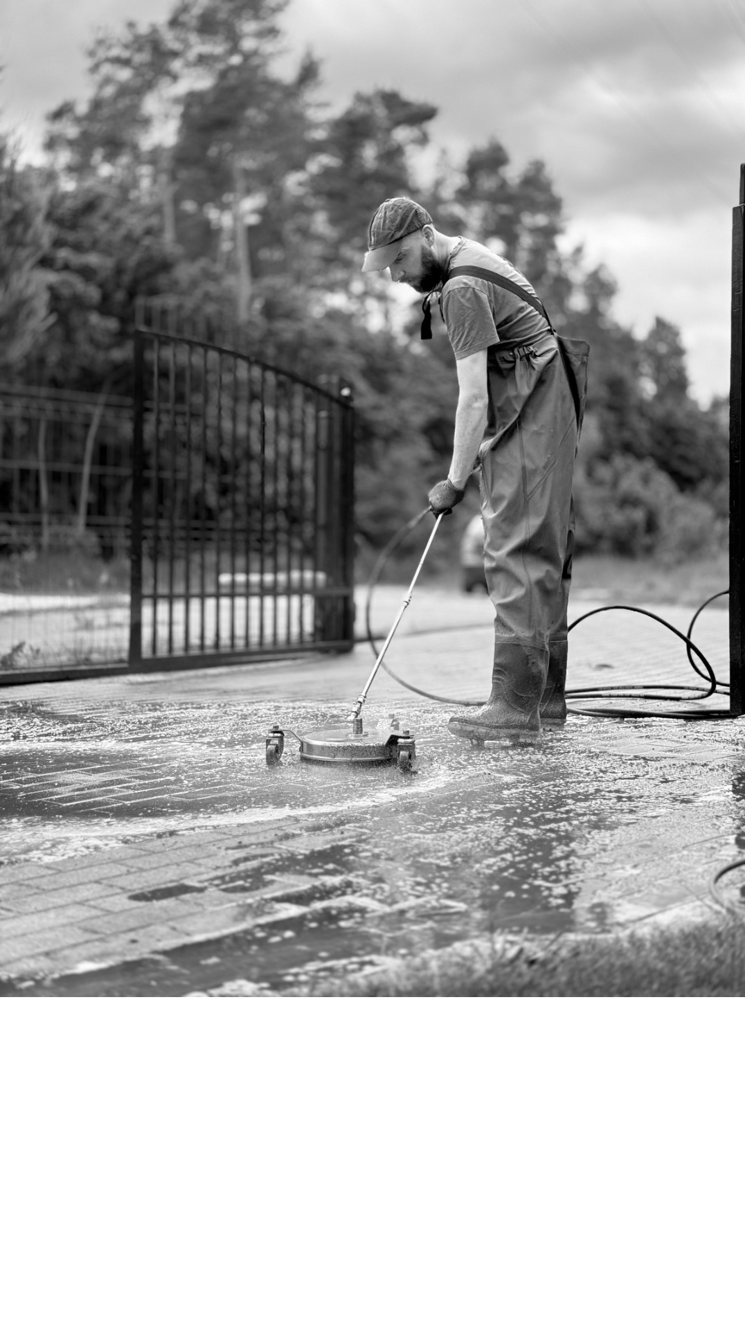 Pracownik w kombinezonie i czapce czyści kostkę brukową za pomocą myjki ciśnieniowej na tle metalowej bramy i zieleni. Czarno-biała fotografia.